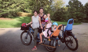 A family of a mother, father, and two kids are standing by their e-bike. The two kids are strapped into the back of the e-bike.