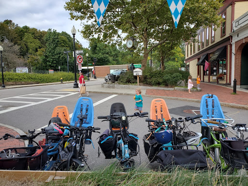 Several ebikes with cjild seats parked on a city street; children play in the background