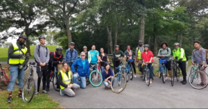 A group of people pose with their bikes in front of a wooded green space