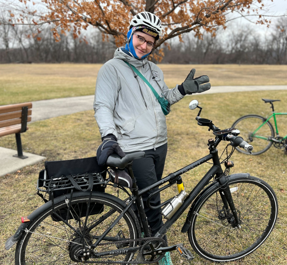 Ryan next to her bike decked out in her winter gear including a nice reflective rain jacket and lobster gloves. She is in a field with fading yellow grass and a pedestrian path.