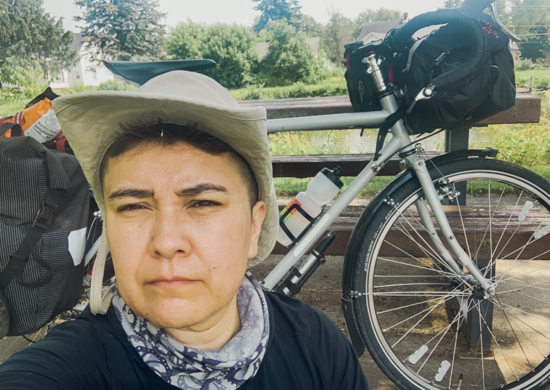 Ari, wearing cowboy hat and standing in front of their Surly Long Haul Trucker bike, loaded with bikepacking gear