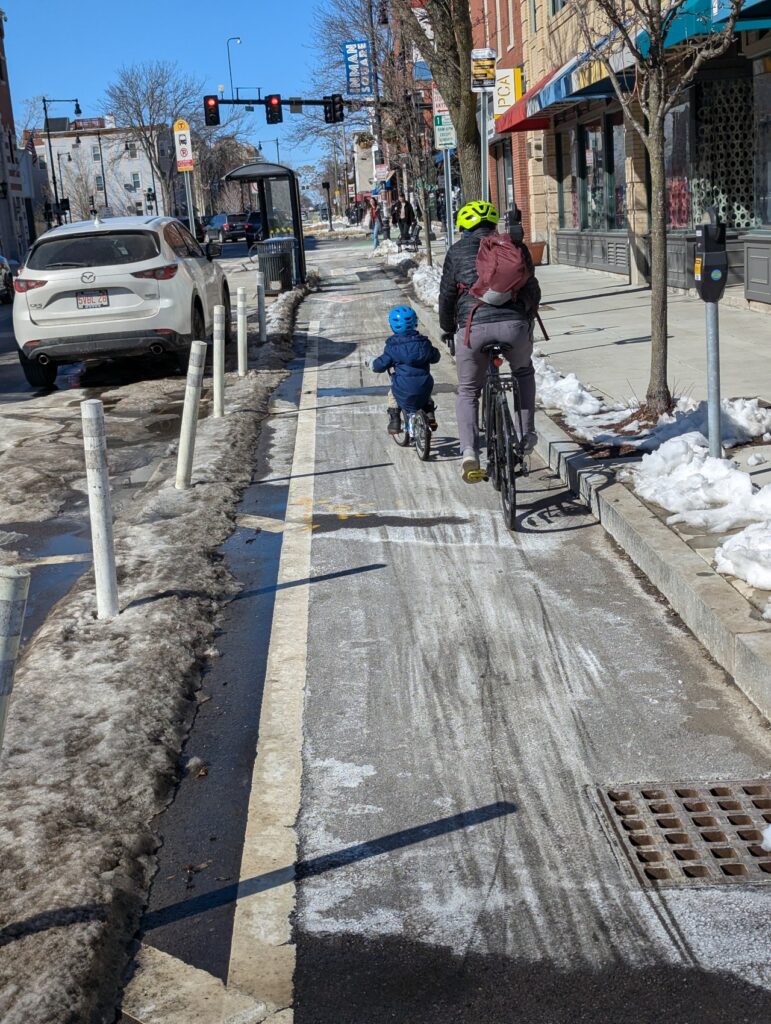 An adult and a child, both wearing coats and helmets, assumed to be a family, is riding through a separated bike lane, paved during the winter.