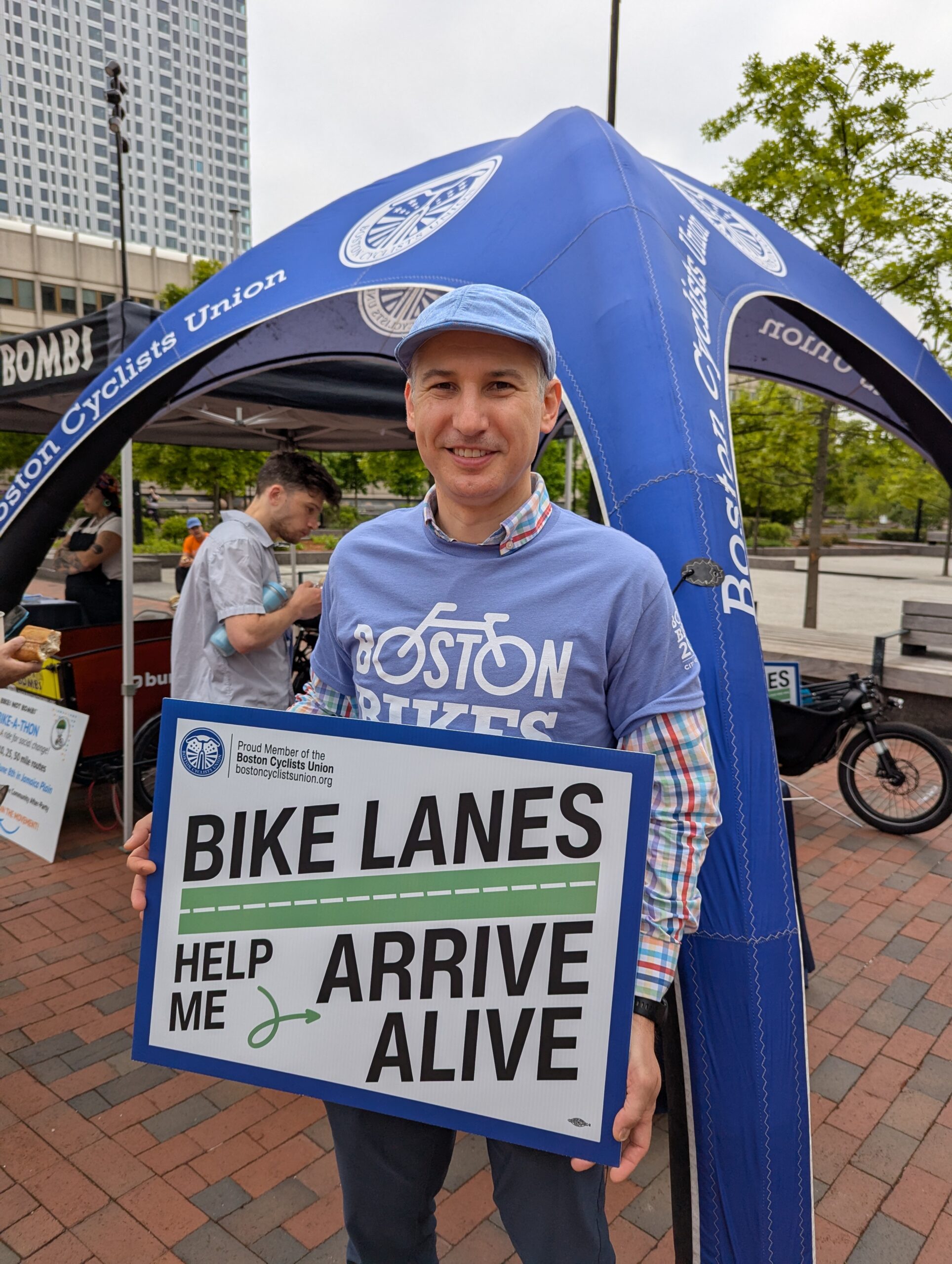 Smiling man in a blue cap and a blue shirt that says "Boston Bikes" holds a yard sign that says "Bike Lanes Help Me Arrive Alive" during a street fair in front of the Boston Cyclists Union tent
