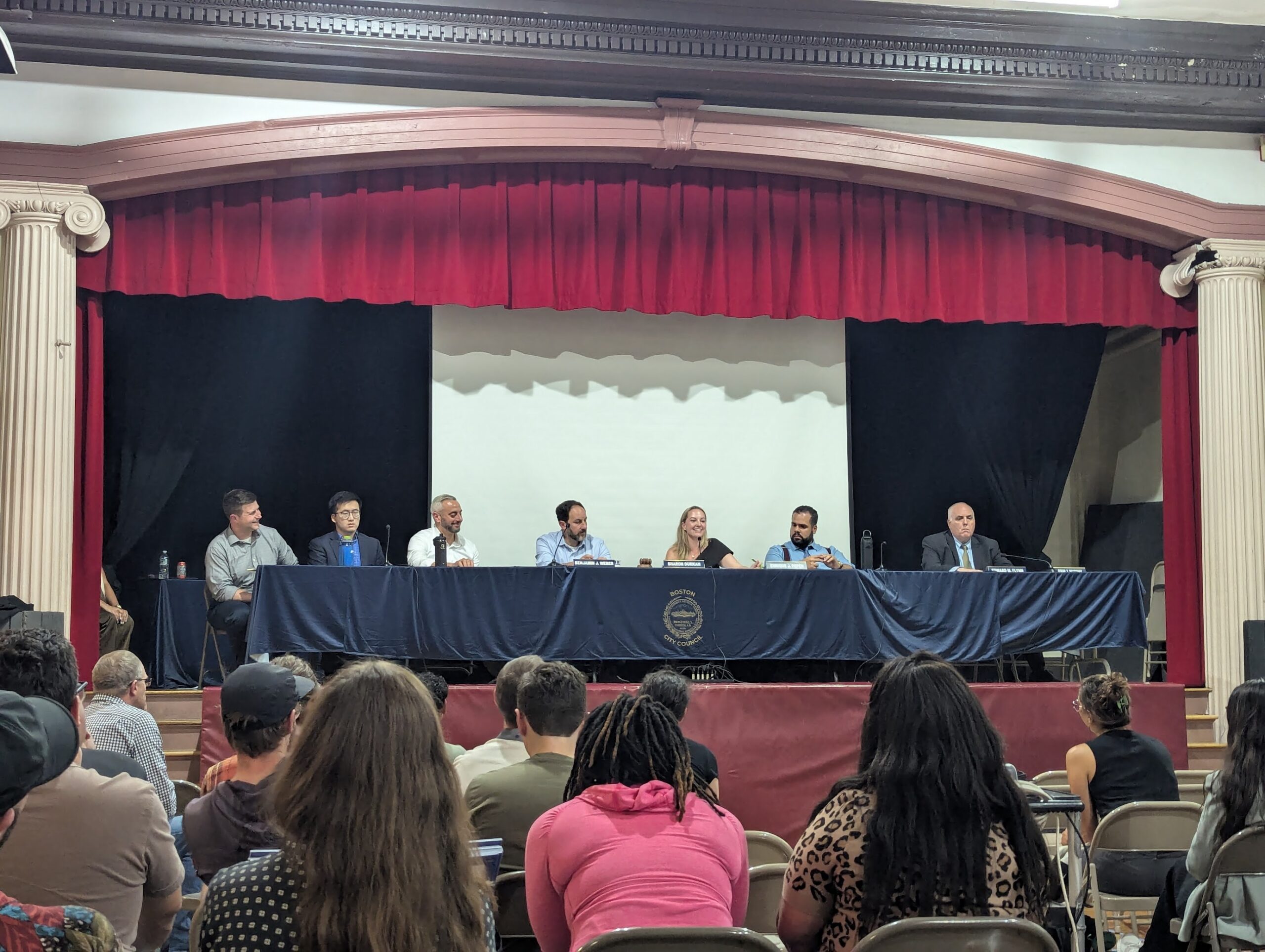 City councilors in charge of the Wednesday October 6th public hearing on Hyde Park Avenue sit on a stage behind a table with a blue city of Boston tablecloth, while a full room sits in the room looking towards them.