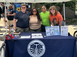Four people stand behind a table with the BCU logo, with BCU materials on it. They're smiling and wearing sunglasses. Anne G, Tiffany, Peter C, and Mandy