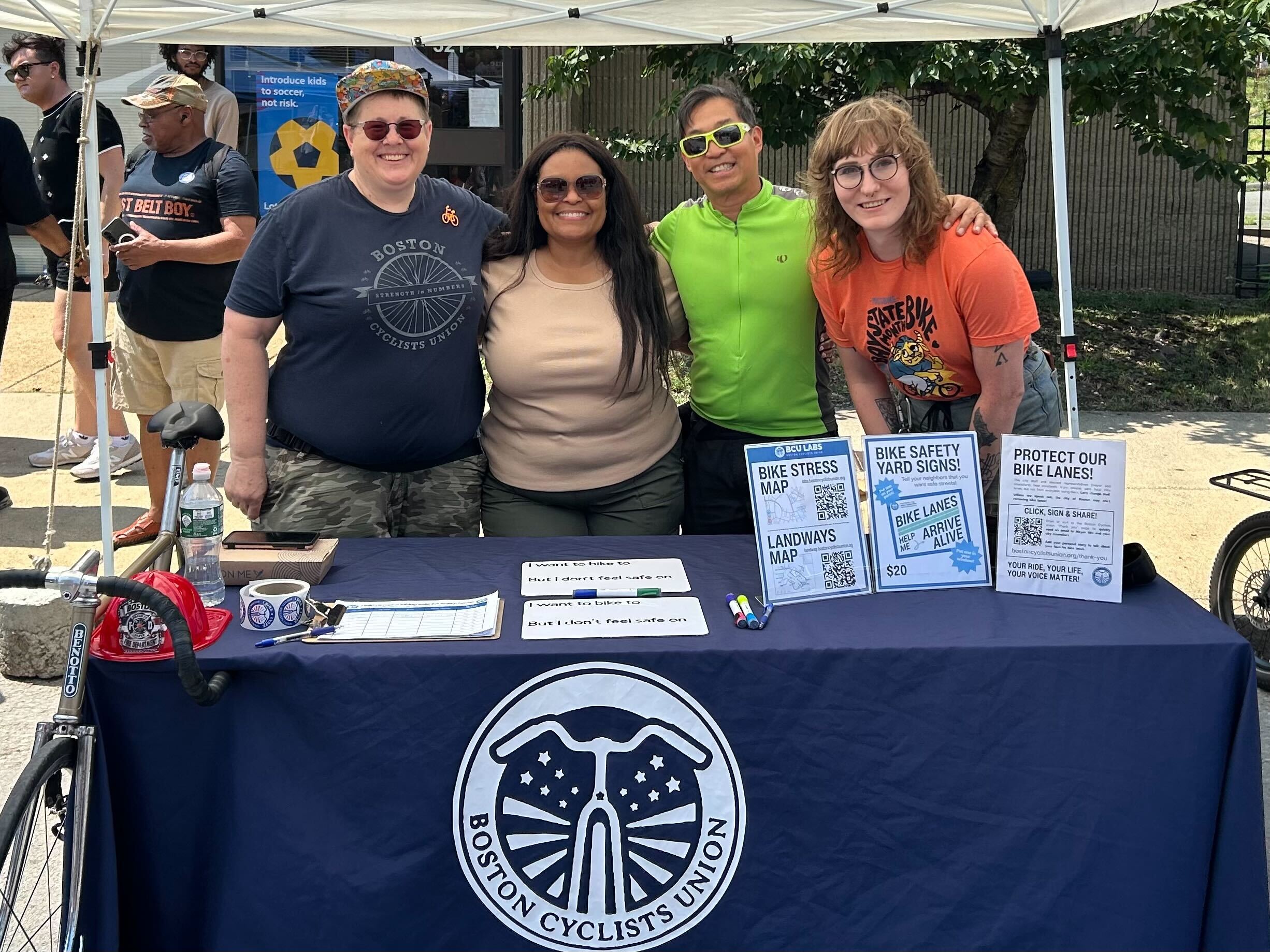 Four people stand behind a table with the BCU logo, with BCU materials on it. They're smiling and wearing sunglasses. Anne G, Tiffany, Peter C, and Mandy