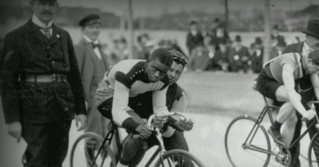 A black man is mounted on his bicycle, held by a coach, ready to get started racing. He looks at the camera with determination. That man is Major Taylor. This is a black and white photograph that's old, likely taken in the late 19th century or early 20th century.