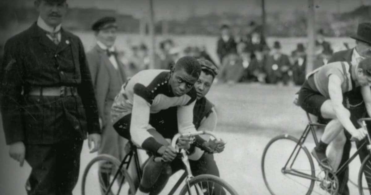 A black man is mounted on his bicycle, held by a coach, ready to get started racing. He looks at the camera with determination. That man is Major Taylor. This is a black and white photograph that's old, likely taken in the late 19th century or early 20th century.