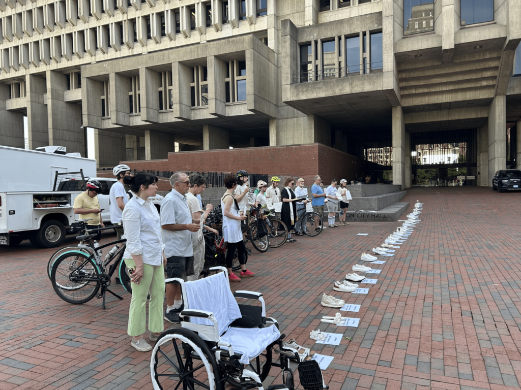 Ride of Silence attendees at City Hall - people dressed in light colored clothing are holding signs behind a row of white shows lined up in the direction of Boston City Hall 